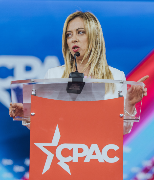 Eine Frau in einem weißen Kleid steht an einem Rednerpult mit einem Mikrofon und sieht aus wie eine Politologin, mit einem unscharfen Hintergrund.