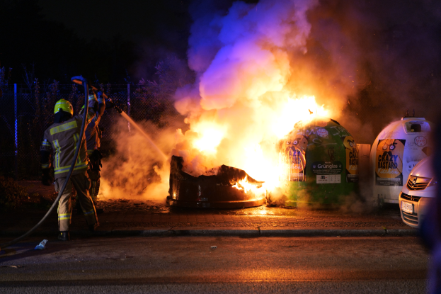 Ein Feuerwehrmann mit Helm und einer Stange steht vor einem brennenden Auto an der Straßenseite, mit anderen Fahrzeugen rechts, einem Zaun, Bäumen und einem dunklen Himmel im Hintergrund.