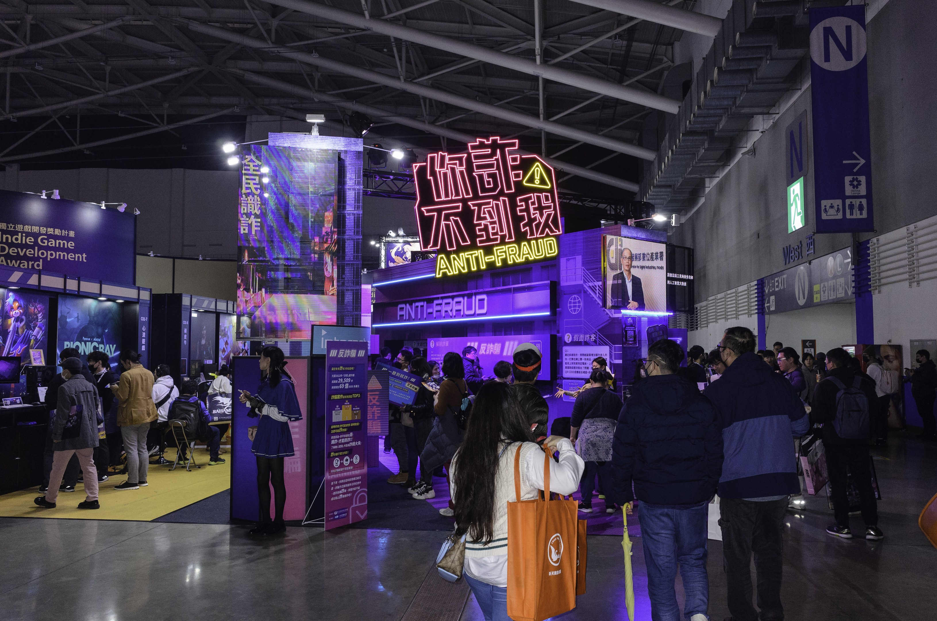 Eine große Gruppe von Menschen steht vor einem Messestand, der den 'Indie Game Development Award 2019' präsentiert, mit Tafeln, Bildschirmen und anderen Gegenständen im Hintergrund.