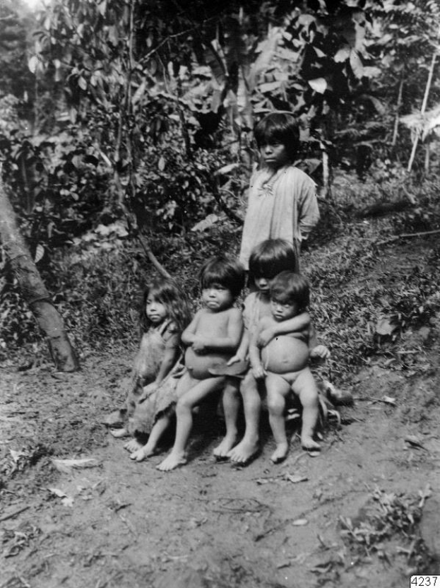 Fünf Kinder sitzen auf einer Schotterstraße in einem Schwarz-Weiß-Bild mit Bäumen und einer stehenden Person im Hintergrund und Text unten; die Kinder scheinen verstört zu sein.
