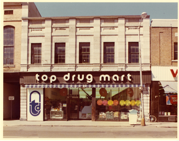 Ein Drogengeschäft an einer Stra√enecke mit einem Gebäude, einem Schild, einem Stra√enpfahl, einem geparkten Fahrrad, einem Müllcontainer und einem bewölktem Himmel.