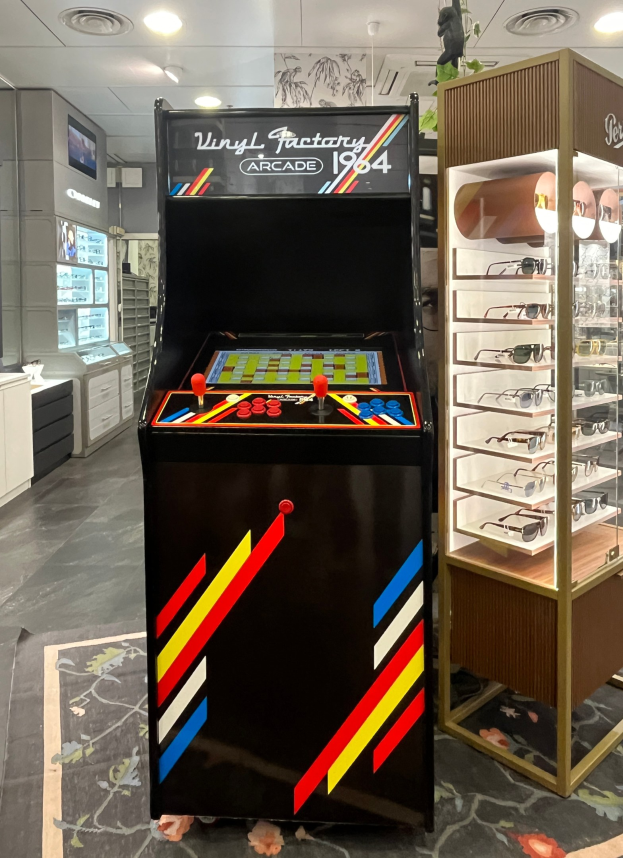 Ein Geschäft mit einem zentralen Retro-Arcade-Automaten, einem rechten Gestell mit Gläsern und Hintergrundelementen wie Schränken, einem Fernseher und anderen Gegenständen, mit Deckenleuchten, die eine Retro-Atmosphäre schaffen.