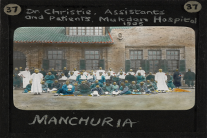 Eine Gruppe von Menschen, die vor einem Gebäude mit Fenstern und Türen auf dem Boden sitzen, mit Text oben und unten, der "Dr. Christie, Assistenten und Patienten, Mukden Hospital, Manchuria" lautet.