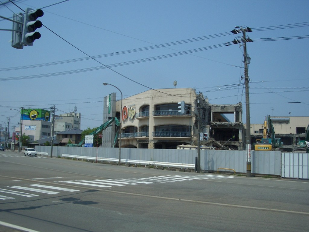 Eine Straßenkreuzung mit Fahrzeugen, Strommasten, Laternen, einer Ampel auf der linken Seite, Gebäuden, Bäumen und dem Himmel im Hintergrund und einem abbruchreifen Gebäude im Vordergrund.