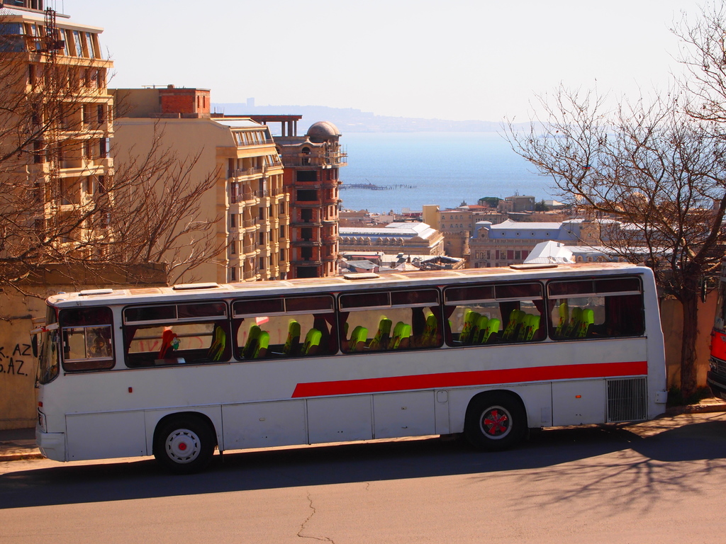 Ein Bus fährt auf der Straße am unteren Bildrand, mit Bäumen und Gebäuden in der Mitte, einem Gewässer auf der rechten Seite und dem Himmel im Hintergrund.
