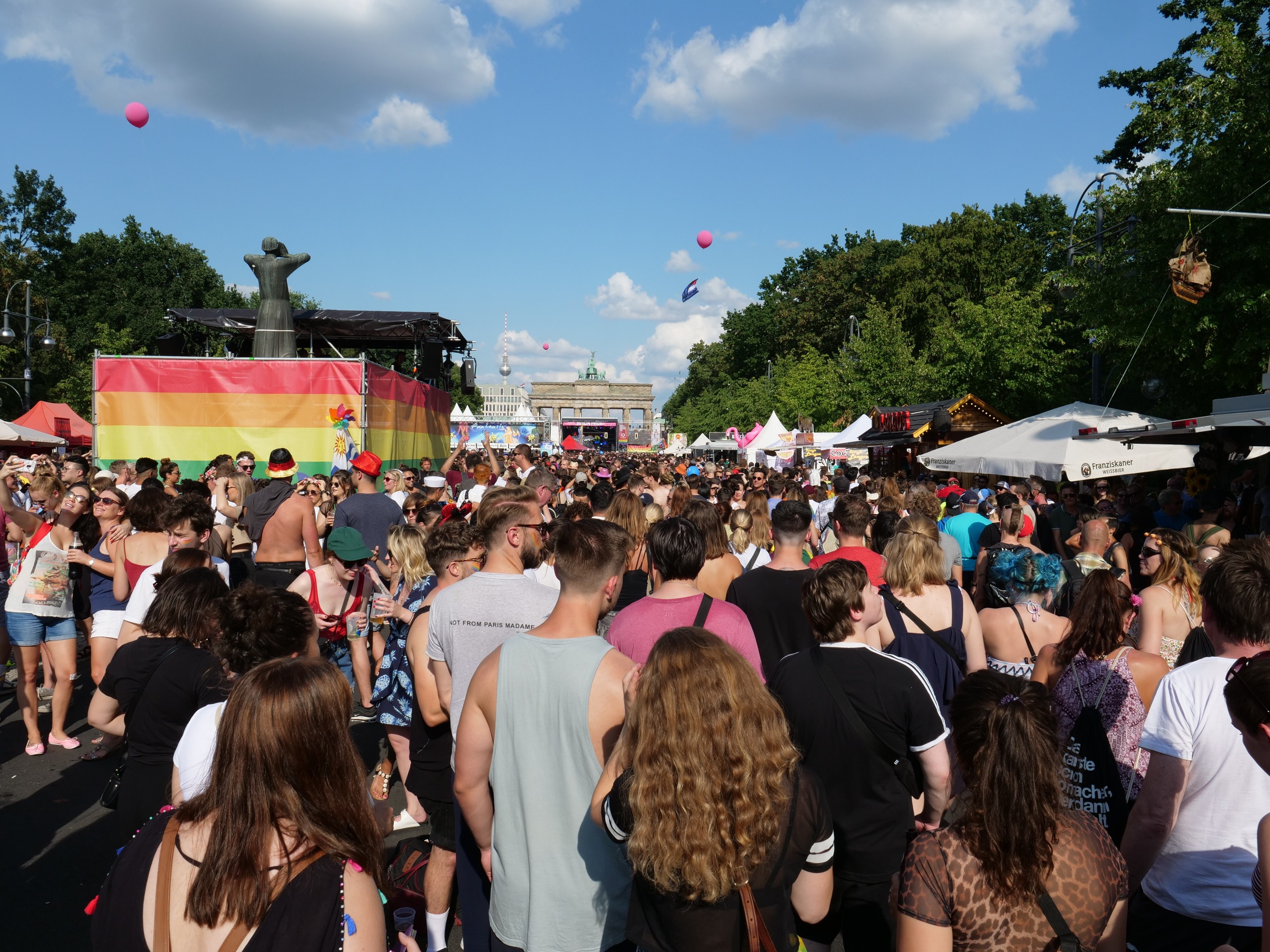 Eine große Menge, die eine Straße mit Zelten, Bäumen, Masten, Lichtern und einer Statue entlanggeht, mit Gebäuden und einem Himmel voller Wolken und Ballons im Hintergrund beim Berlin Pride Festival.