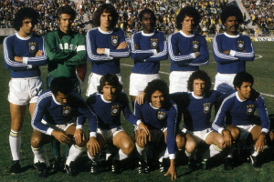 Eine Gruppe von Männern steht auf einem Fußballfeld, einige in einer Hockposition, mit Schildern, die "Brasiliens WM-Mannschaft" und eine Menschenmenge im Hintergrund zeigen.