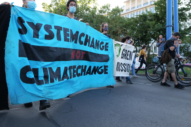 Eine Gruppe von Menschen marschiert mit einem 'System Change Not Climate Change'-Schild die Straße entlang, einige tragen Masken, mit Fahrrädern in der Nähe und Gebäuden, Bäumen und einem klaren blauen Himmel im Hintergrund.