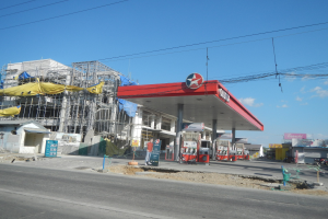 Tankstelle im Bau neben einer Straße mit Gebäuden im Hintergrund, Fahrzeuge vorhanden, verstreute Baustoffe, Masten und Überlandleitungen gegen einen bewölkten Himmel.