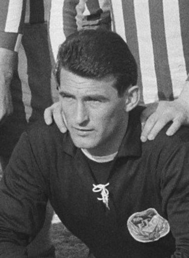 Schwarzes und weißes Foto eines Mannes, der auf einem Rasenfüüballfeld sitzt und ein T-Shirt und eine Kette trägt, mit zwei Personen hinter ihm.