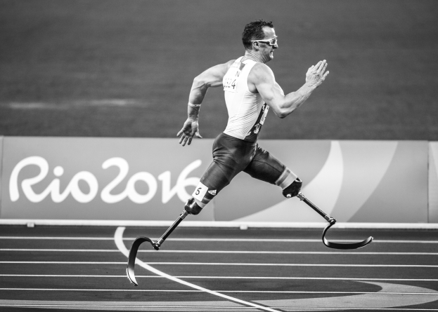 Schwarzes und weißes Bild eines Mannes mit einem Prothesenbein, der auf einer Laufbahn an den Rio 2016 Paralympischen Spielen läuft, mit dem Namen des Ereignisses auf einer Tafel im Hintergrund.