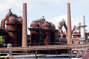 Großes Industriegebäude mit ausgedehnten Rohrleitungen, mehreren Schornsteinen, einer Brücke, Strommasten mit Lampen, umliegenden Bäumen und einem bewölkten Himmel.