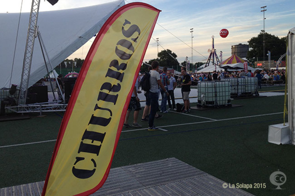 Ein buntes Karnevalszelt mit Zelten, einem zentralen Mast, Menschen, Bäumen, Strommasten, Straßenbahnen auf der Straße und einem Eisenständer auf der linken Seite unter einem klaren Himmel.