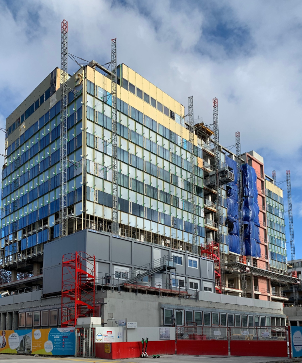 Großes Gebäude im Bau in einer Stadt mit Glasfenstern, Metallrahmen, einer Treppe, einem Zaun, einem Schild, Pfosten, einer Straßenlaterne, Bäumen und einem bewölkten Himmel.