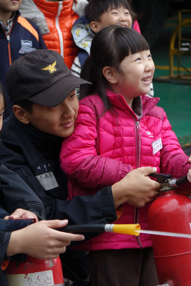 Gruppe von Kindern um einen roten Hydranten in einer Schulumgebung, ein Kind hält ein Rohr.