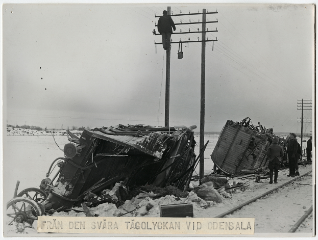 Ein Schwarz-Weiß-Foto von einer Zugkatastrophe auf den Schienen mit ein paar Leuten, die rechts daneben stehen, Masten, Drähten, Bäumen und dem Himmel im Hintergrund und Text unten.