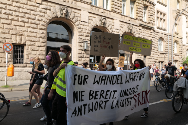 Eine Gruppe von Menschen marschiert auf der Straße in Berlin, hält Schilder und Banner hoch und fährt mit Fahrrädern vorbei an einem Gebäude mit Bögen, Säulen, Skulpturen und Bäumen.