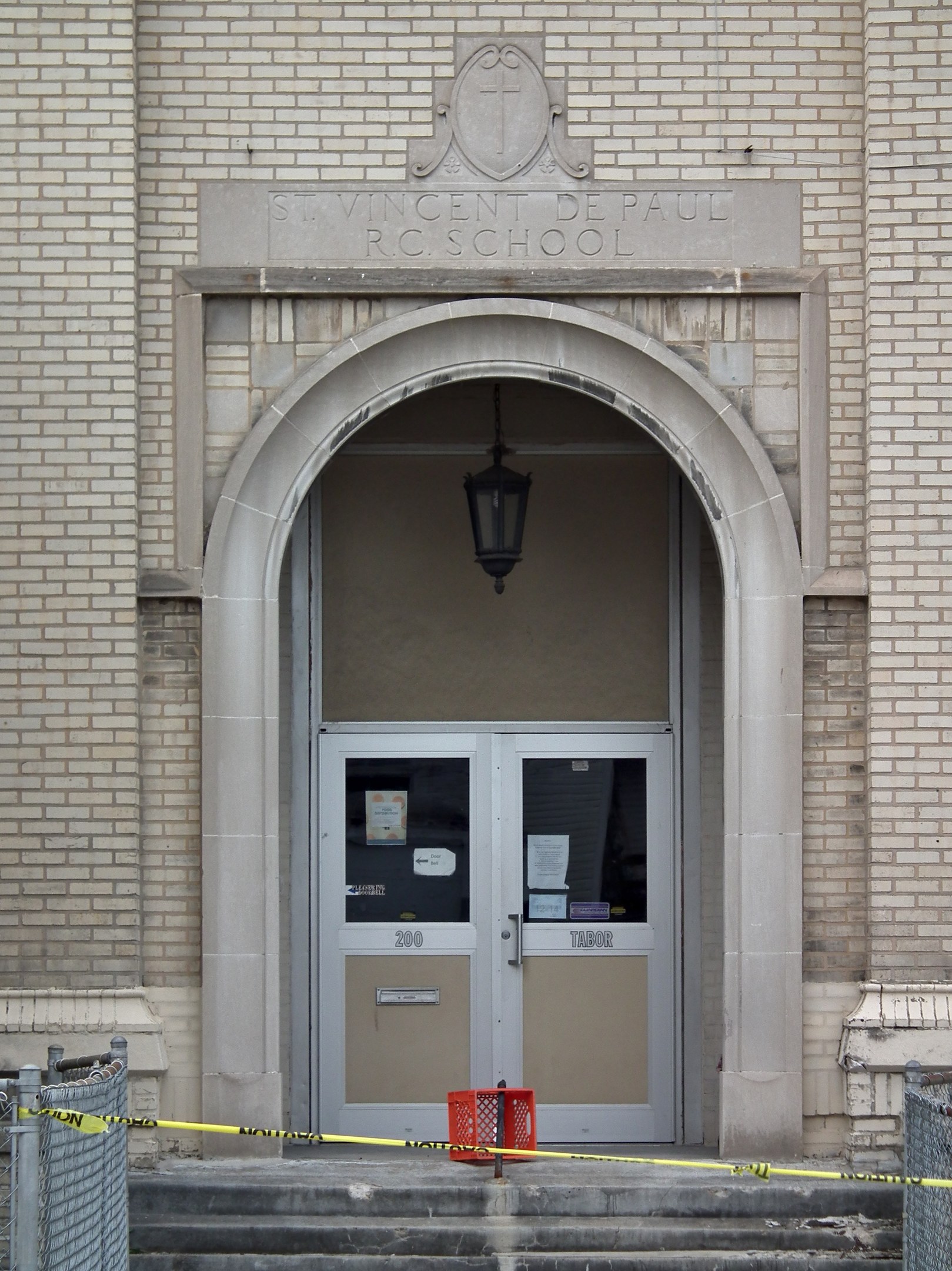 Ein Eingang eines Backsteingebäudes mit gelbem Absperrband, Treppen mit Maschendrahtzaun, eine Tür mit Papieren, eine hängende Lampe und eine Wandinschrift, die wahrscheinlich den Namen der Schule angibt.
