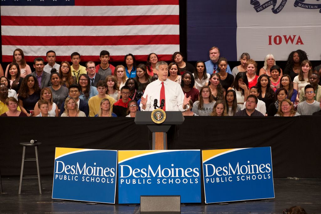 Ein Mann in weißem Hemd und roter Krawatte steht an einem Rednerpult mit drei Tafeln mit Text, einem Hocker mit Mikrofon und einer sitzenden Zuschauermenge im Hintergrund, sowie einem Flaggenmuster und zusätzlichem Text.