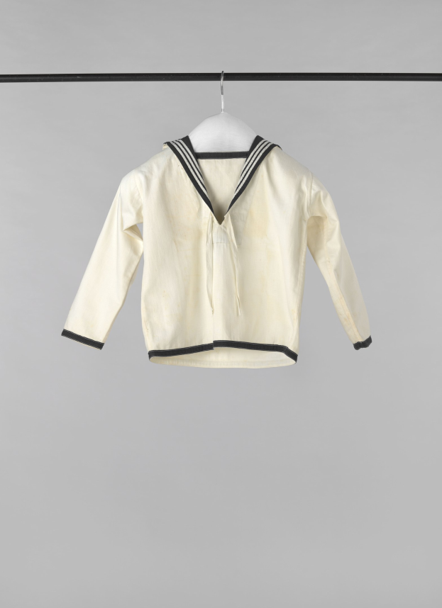 Eine weiße Matrosenjacke mit Kragen, langen Ärmeln und Knopfleiste, die an einer Wäscheleine gegen einen weißen Hintergrund hängt.