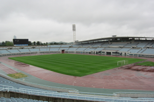 Großes Stadion mit einem Fußballfeld, umgeben von Sitzplätzen, einem Zaun, einem Display, Bäumen, einem Turm, Gebäuden und einem bewölkten Himmel.
