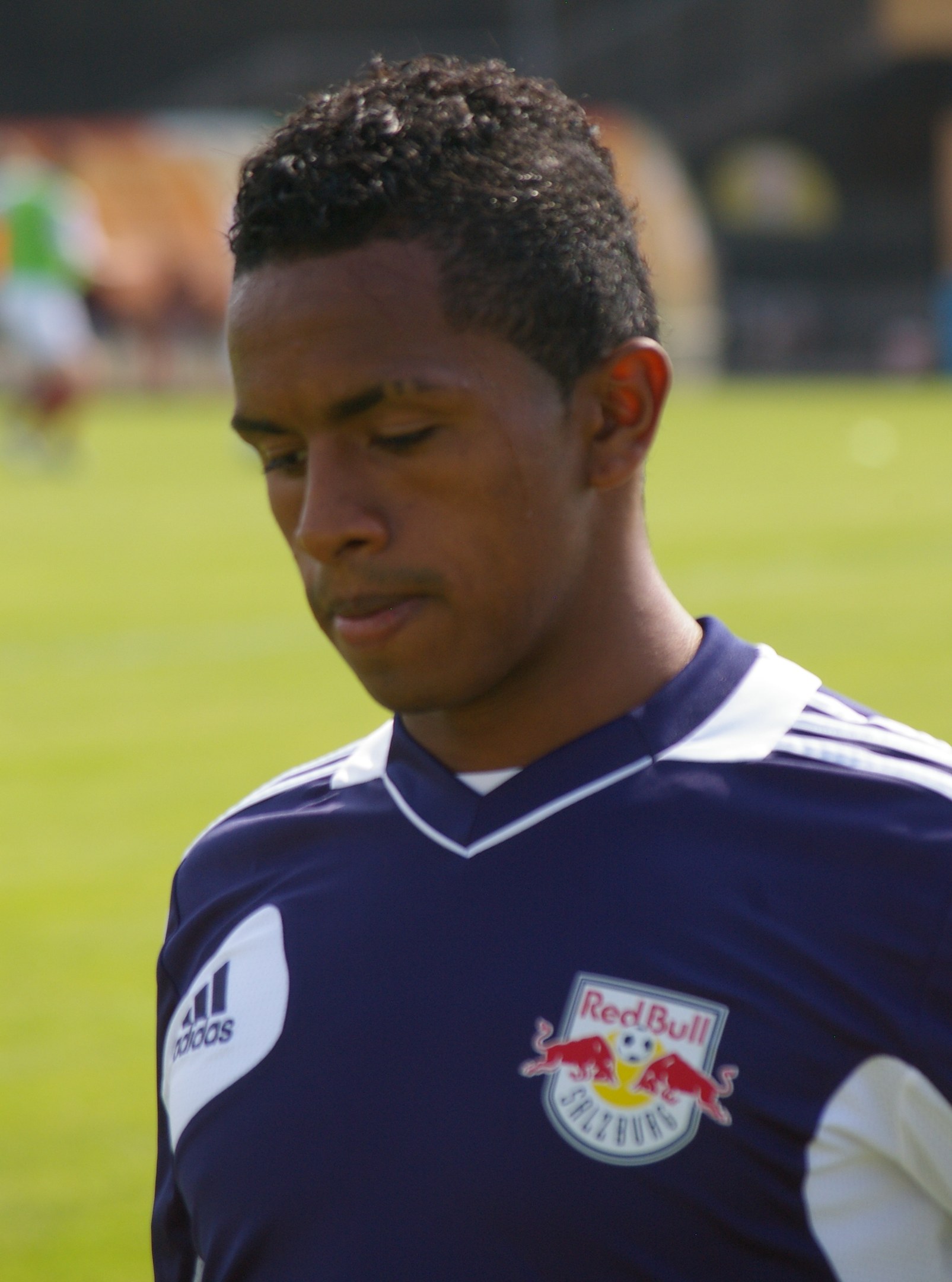 Junger Mann in blauem T-Shirt auf einem Fußballfeld stehend, der als Flügelspieler mit kurzen dunklen Haaren und entschlossener Miene erscheint, vor einem unscharfen Hintergrund.