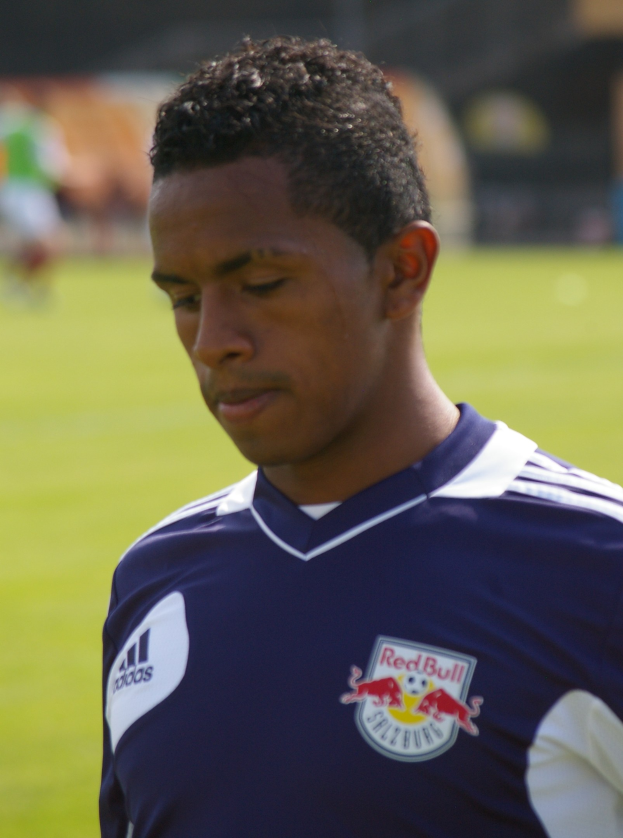 Junger Mann in blauem T-Shirt auf einem Fußballfeld stehend, der als Flügelspieler mit kurzen dunklen Haaren und entschlossener Miene erscheint, vor einem unscharfen Hintergrund.