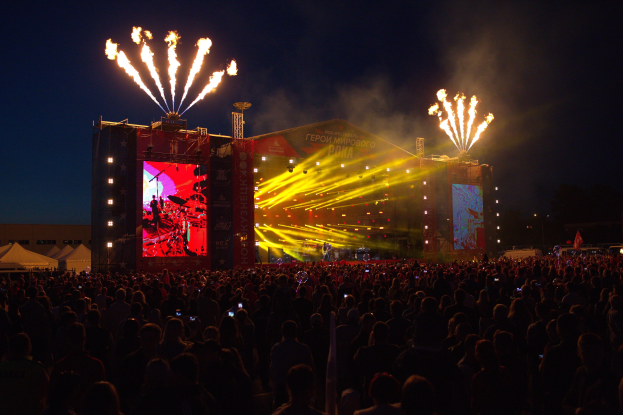 Eine große Menschenmenge auf einem Musikfestival schaut Zuschauern auf einer hell erleuchteten Bühne mit Bannern, umgeben von Zelten und Feuerwerk am dunklen Himmel.