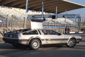 Ein silberner DeLorean DMC-12 fährt auf einer Straße vorbei an Menschen, Fahnen, Schildern, Geländern, Stühlen und einer Hütte, mit Bäumen und einem klaren blauen Himmel im Hintergrund.