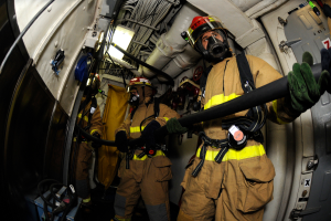 Eine Gruppe von Feuerwehrmännern in Helmen und Handschuhen hält einen Schlauch am Heck eines Flugzeugs, mit Kabeln, Lampen und anderen Gegenständen im Hintergrund und Text am unteren Bildrand.