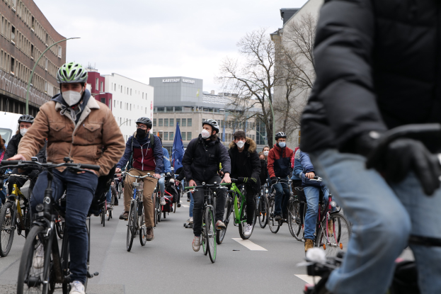 Eine Gruppe von Menschen in Helmen und Handschuhen fährt mit Fahrrädern eine von Bäumen gesäumte Straße in Berlin, Deutschland, entlang, wobei Gebäude und ein geparktes Fahrzeug im Hintergrund zu sehen sind.