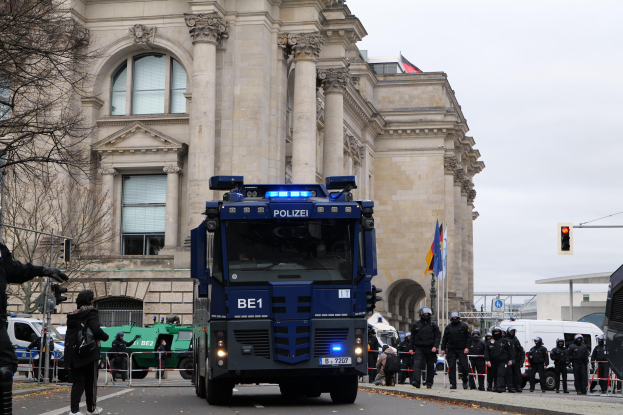 Eine Gruppe von Polizisten steht vor einem großen Gebäude mit Fenstern, Säulen und Bögen, mit Fahrzeugen auf der Straße, einer Person mit einer Kamera auf der linken Seite und einem klaren blauen Himmel im Hintergrund.