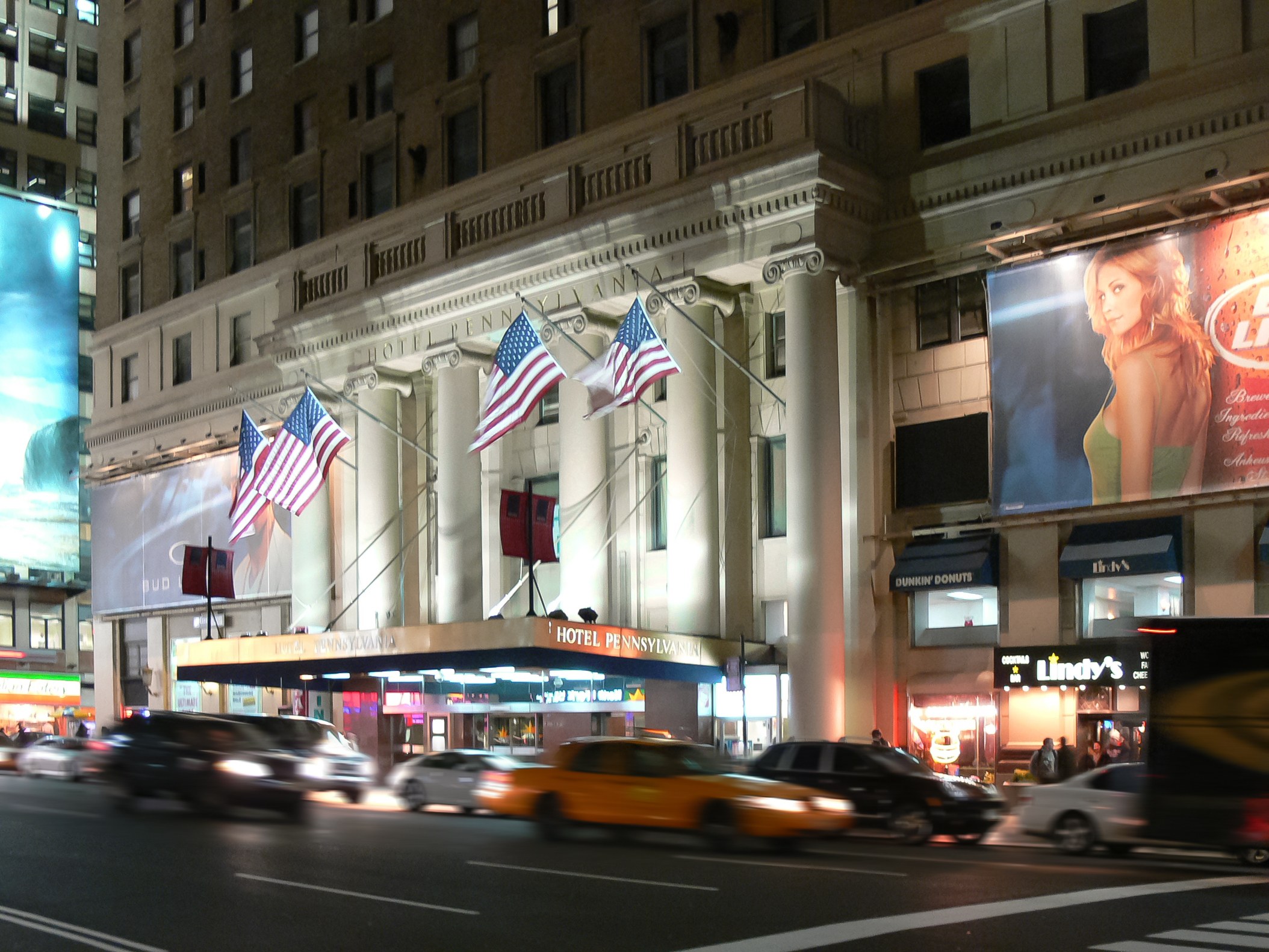 Belebte Stadtstraße bei Nacht mit fahrenden Fahrzeugen, einem Gebäude mit Fenstern, Säulen und Flaggen im Hintergrund, beleuchtete Schilder mit Text und einem Bild einer Frau sowie Straßenbeleuchtung.
