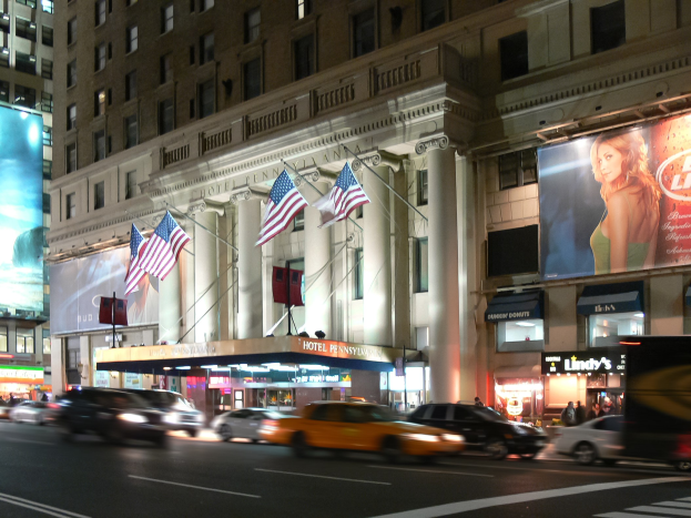 Belebte Stadtstraße bei Nacht mit fahrenden Fahrzeugen, einem Gebäude mit Fenstern, Säulen und Flaggen im Hintergrund, beleuchtete Schilder mit Text und einem Bild einer Frau sowie Straßenbeleuchtung.