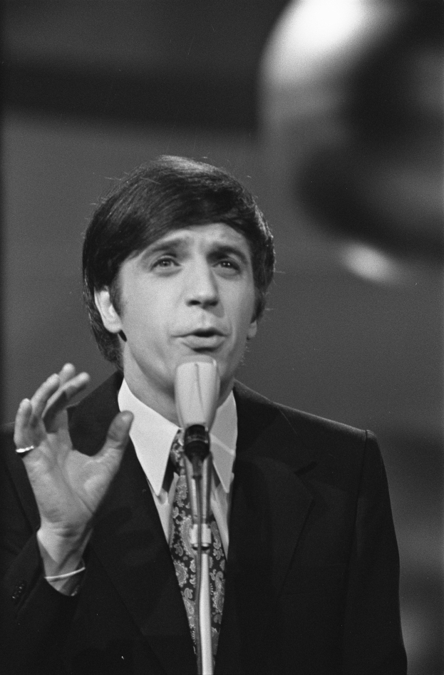 Ein Mann in einem schwarzen Anzug, weißem Hemd und gemustertem Schlips spricht ernst in ein Mikrofon, sein Haar ist zurückgekämmt.