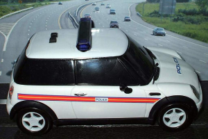 Ein weißes Polizeiauto steht im Vordergrund, dahinter fahren mehrere Autos auf der Straße, flankiert von Gras und Bäumen auf beiden Seiten in einer ländlichen Umgebung.