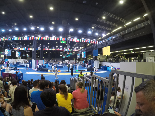 Eine große Menschenmenge sitzt in einer Indoor-Sportarena vor einer Bühne, mit Stehplätzen auf dem Boden, sichtbaren Geländern, Bildschirmen, Flaggen und Deckenleuchten.