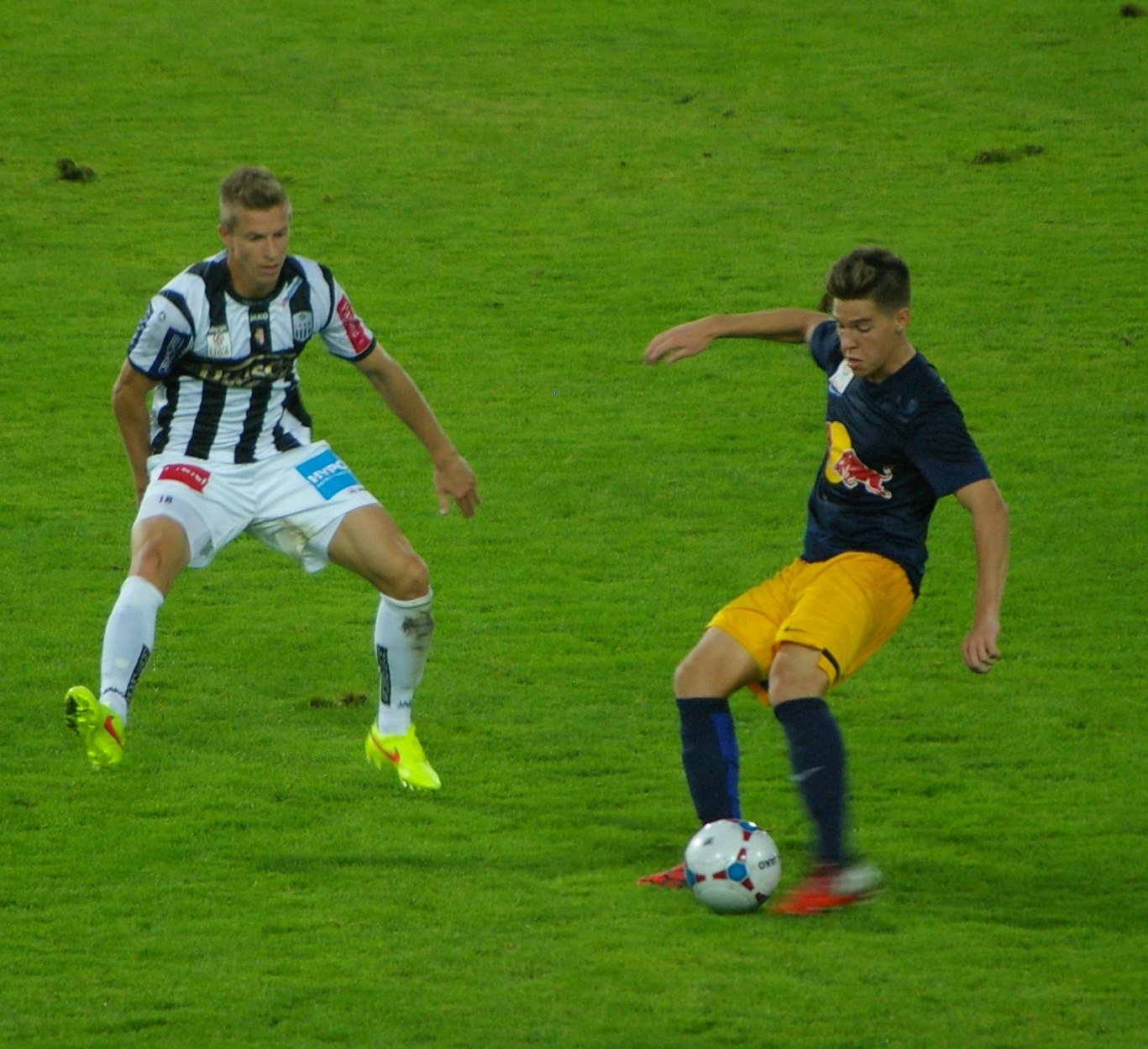 Zwei Männer spielen Fußball auf einem grünen Feld, einer trägt ein blaues T-Shirt und gelbe Shorts, der andere ein weißes und schwarzes gestreiftes T-Shirt und weiße Shorts.