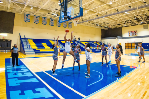Eine Gruppe von Frauen, die Basketball in einer Turnhalle mit einem Scoreboard spielt, das ihren Sieg in der NCAA-Meisterschaft anzeigt, Zuschauer auf den Tribünen und Banner an der Wand.