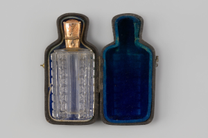 Eine gläserne Parfümflasche mit zylindrischer Form und engem Hals, die in einem tiefblauen Etui mit goldener Verzierung auf einer weißen Oberfläche steht.