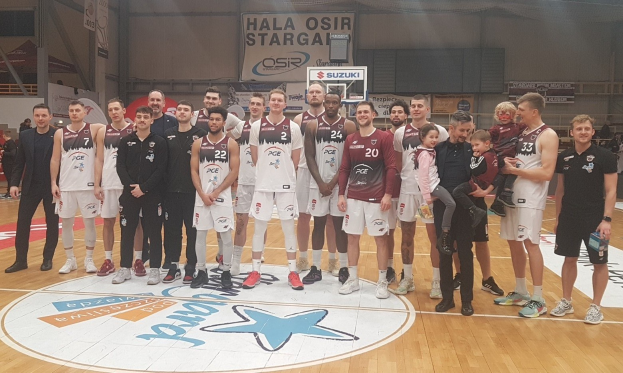 Männer- und Kinder-Gruppe auf einem Basketballfeld mit Schildern "Hala Osir Starga", Zuschauer und Equipment im Hintergrund.