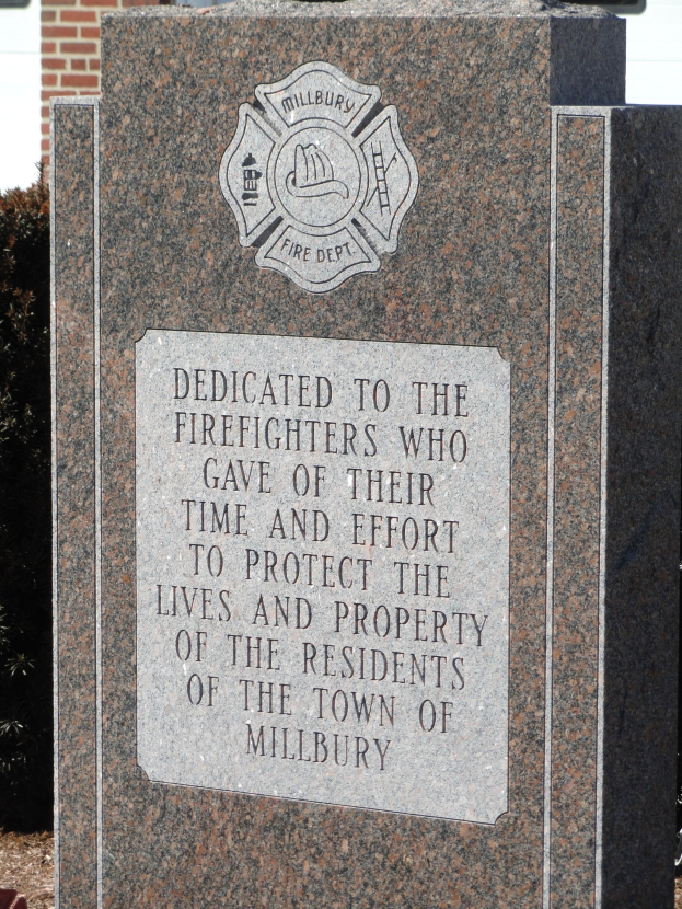 Denkmalstein mit einer Gedenktafel für Feuerwehrleute, vor einem Backsteingebäude mit umliegenden Pflanzen und Bäumen gesetzt.