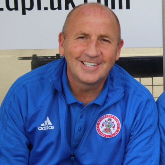Zwei Männer in einem Raum, einer in einem blauen Jackett, der neben einem anderen Mann lächelt, mit einer Tafel an der Wand im Hintergrund.