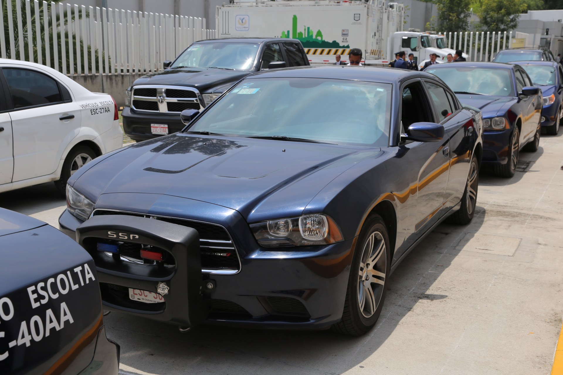 Eine Gruppe von Polizeiwagen nebeneinander auf einem Parkplatz geparkt, mit Menschen auf der Straße im Vordergrund, ein Zaun, Bäume, Gebäude und einen klaren blauen Himmel im Hintergrund.