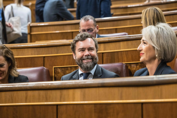 Gruppe von Menschen, die in einem Gerichtssaal sitzen, mit einem Mann in Anzug und Krawatte und zwei Frauen in legerer Kleidung nebeneinander, Bänke im Hintergrund.