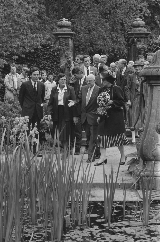 Schwarzes und weißes Bild einer Gruppe von Menschen, die sich um einen Teich in einem Garten versammelt haben, mit einer Frau in der Mitte, die einen Blumenstrauß hält, und einem Mann in einem Anzug rechts von ihr, umgeben von üppigen Pflanzen, Bäumen und Säulen im Hintergrund.
