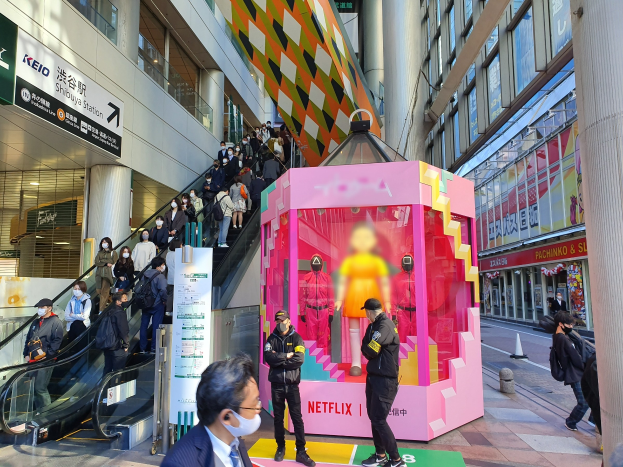 Eine Gruppe von Menschen steht vor einem Gebäude mit Glaswänden und Texttafeln, einige tragen Masken, mit einer Rolltreppe im Hintergrund; eine pinkfarbene Box in der Mitte zeigt Schaufensterpuppen und trägt den Text "Netflix' neue Netflix-Bühne in Tokio."