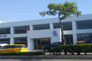 Ein Volkswagen-Autohaus mit parkenden Autos, umgeben von Pflanzen und einem Baum, mit einem gläsernen Gebäude und einem sichtbaren Himmel im Hintergrund.