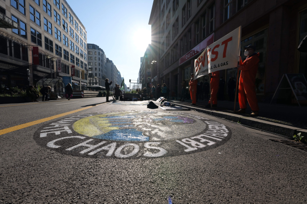 Eine Gruppe von Menschen mit Schildern und Plakaten auf einer Stadtstraße während einer Demonstration, mit Gebäuden, Pflanzen und einer gemalten "Chaos"-Nachricht auf der Straße.