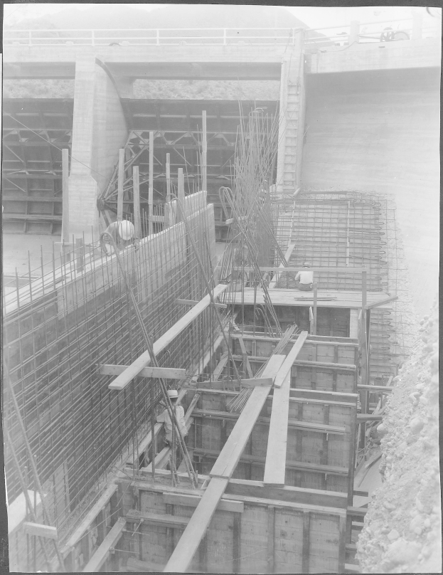 Ein Schwarz-Weiß-Foto einer Baustelle mit einer Brücke im Hintergrund, das Arbeiter, Stangen, hölzerne Gegenstände und andere Baustoffe zeigt, mit Text unten.